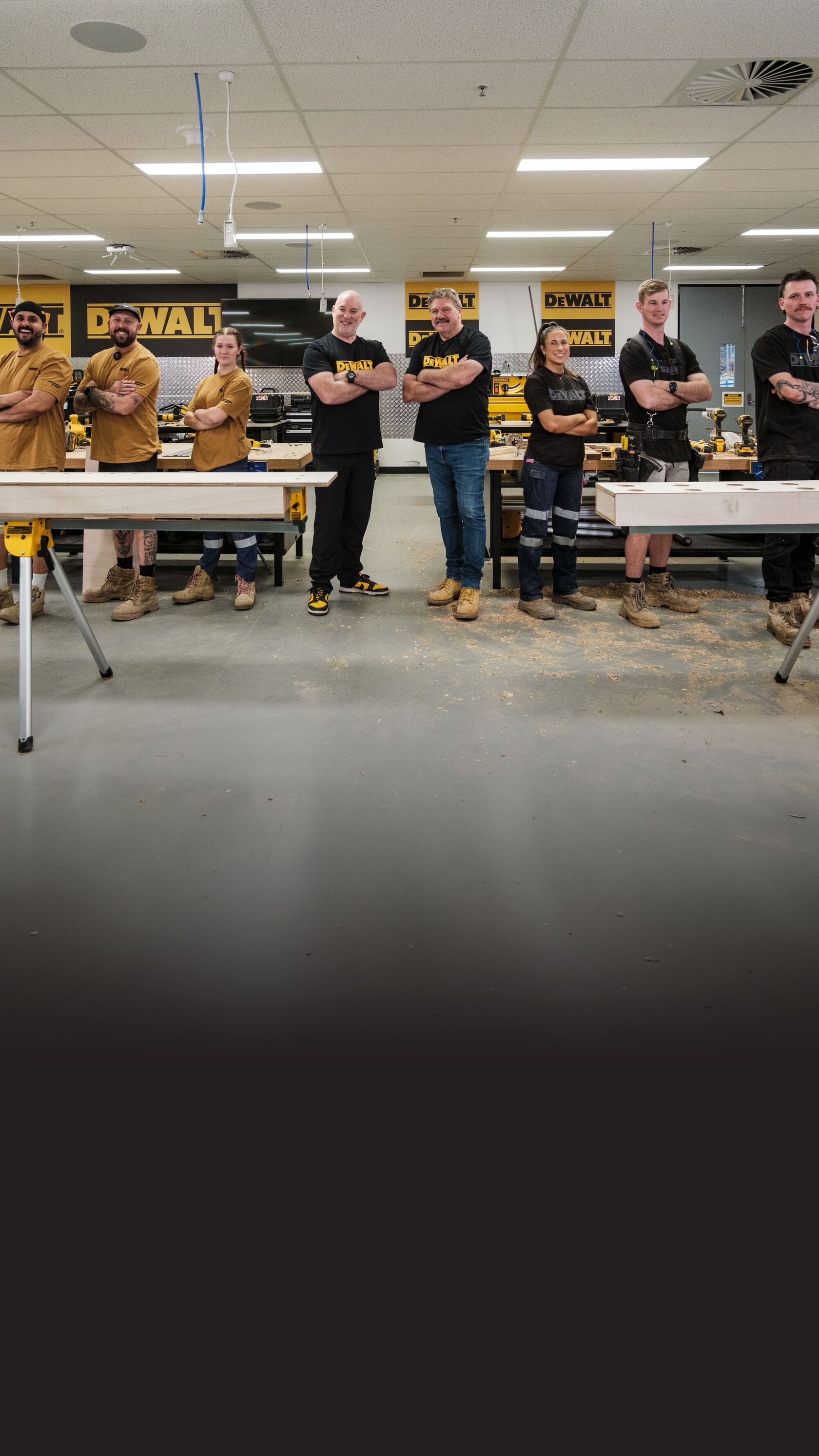 Group of people in work attire standing in a DEWALT workshop, posing with arms crossed near workbenches.