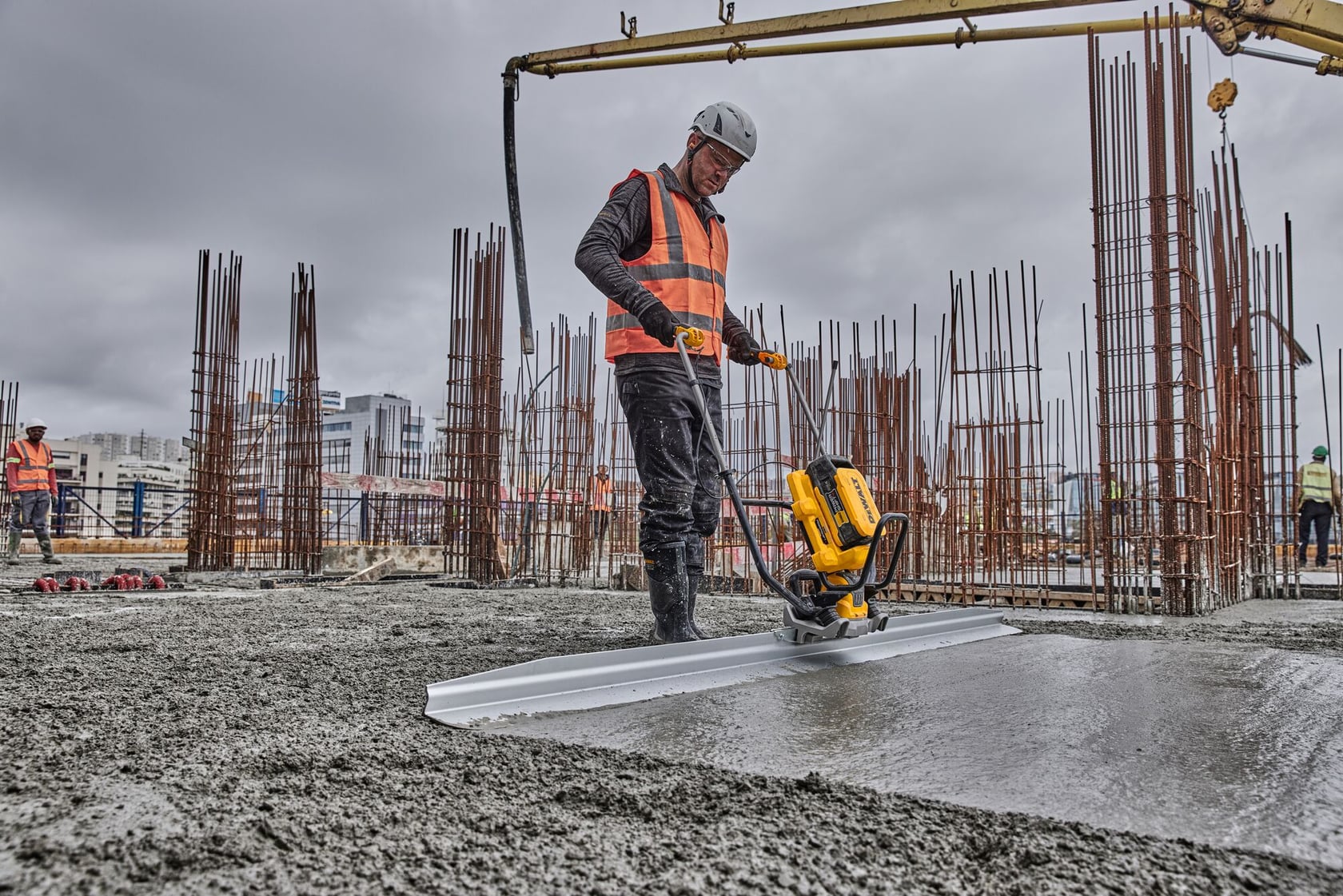 A construction worker wearing a safety vest and helmet uses a DEWALT power tool to smooth wet concrete on a building site. Reinforced steel bars and construction workers are visible in the background under a cloudy sky.